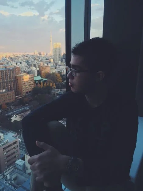A solo Gen Z individual sitting by a window, looking out at a city skyline, with a thoughtful expression, symbolizing reflection on purpose and future. Soft, introspective lighting.