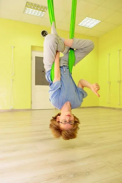 A joyful child swinging on a suspension swing in a brightly lit sensory gym, with soft mats on the floor, indicating controlled and safe sensory play.