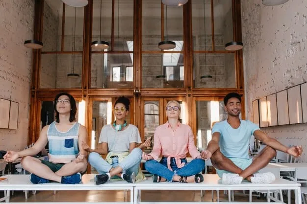 A diverse group of Gen Z students seated at desks, practicing mindful breathing with their hands on their stomachs, guided by a teacher standing calmly at the front of the classroom.