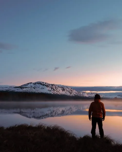 A lone Gen Z hiker gazing out at the dramatic, untamed landscape of Patagonia, with jagged mountain peaks, a vast glacial lake, and expansive untouched wilderness in the foreground during sunset.