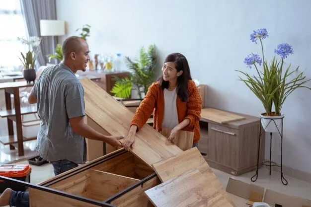 An older woman and a young man are sharing a laugh while assembling furniture in a comfortable, bright living room, symbolizing collaborative intergenerational activity and shared positive experiences.
