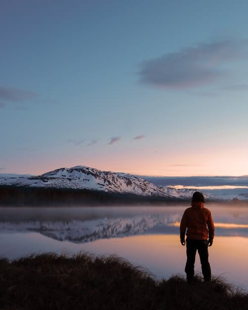 A lone Gen Z hiker gazing out at the dramatic, untamed landscape of Patagonia, with jagged mountain peaks, a vast glacial lake, and expansive untouched wilderness in the foreground during sunset.