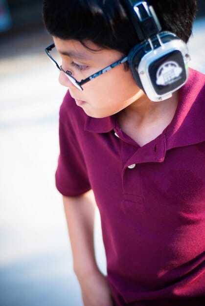 A calm and focused young student, perhaps 9-11 years old, wearing headphones and using a tablet to read along with an audiobook in a classroom setting. The background is slightly blurred but suggests a supportive learning environment.