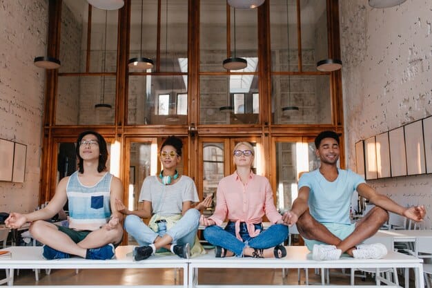 A diverse group of Gen Z students seated at desks, practicing mindful breathing with their hands on their stomachs, guided by a teacher standing calmly at the front of the classroom.