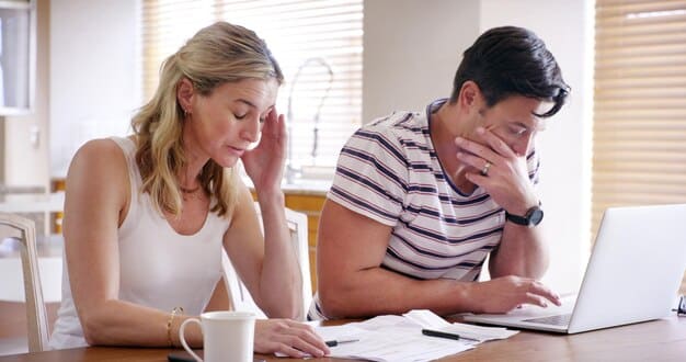 A young couple looking stressed while reviewing mortgage documents in front of a computer, with student loan statements visible on the table. Their faces convey financial burden and uncertainty about future stability.