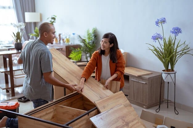 An older woman and a young man are sharing a laugh while assembling furniture in a comfortable, bright living room, symbolizing collaborative intergenerational activity and shared positive experiences.