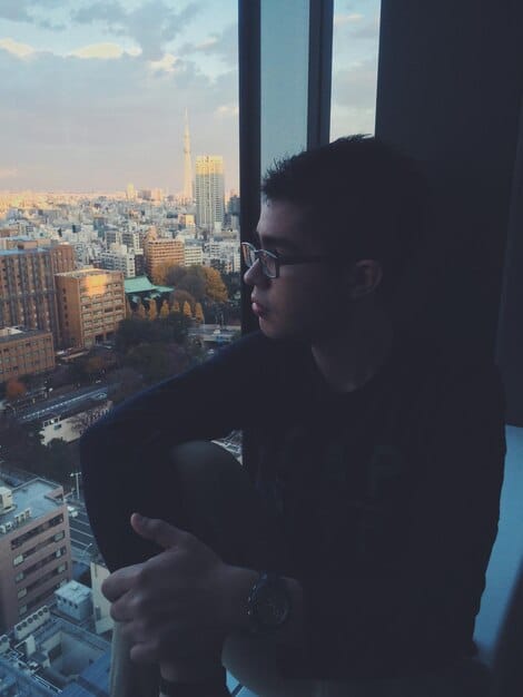 A solo Gen Z individual sitting by a window, looking out at a city skyline, with a thoughtful expression, symbolizing reflection on purpose and future. Soft, introspective lighting.