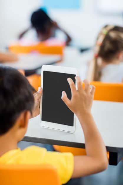 A close-up of a student's hand choosing options from a tablet screen during a classroom activity, symbolizing autonomy in learning. The background is slightly blurred but suggests a classroom setting.